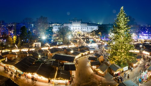 Blick auf den Wiener Weihnachtsmarkt bei Nacht. Im Hintergrund sind historische Gebäude, im Vordergrund sind Weihnachtsmarkthütten und ein festlich beleuchteter Tannenbaum.