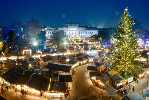 Blick auf den Wiener Weihnachtsmarkt bei Nacht. Im Hintergrund sind historische Gebäude, im Vordergrund sind Weihnachtsmarkthütten und ein festlich beleuchteter Tannenbaum.