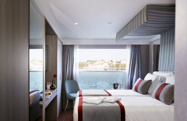 Eine stilvoll eingerichtete Kabine auf der A-ROSA ALVA mit Doppelbett, Schreibtisch und bodentiefem Panoramafenster bei Tageslicht.