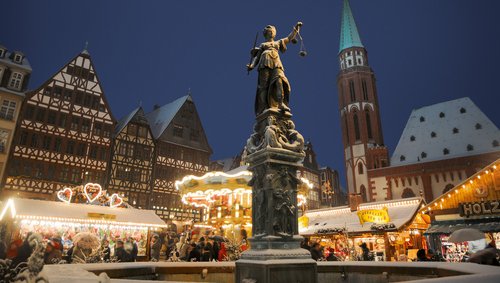 Weihnachtsmarkt in Frankfurt bei Nacht mit dem Justitia-Brunnen im Vordergrund. Festlich beleuchtete Stände und viele Menschen, umgeben von historischen Gebäuden unter einem klaren dunklen Himmel.