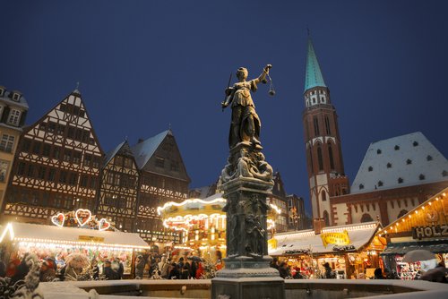 Weihnachtsmarkt in Frankfurt bei Nacht mit dem Justitia-Brunnen im Vordergrund. Festlich beleuchtete Stände und viele Menschen, umgeben von historischen Gebäuden unter einem klaren dunklen Himmel.