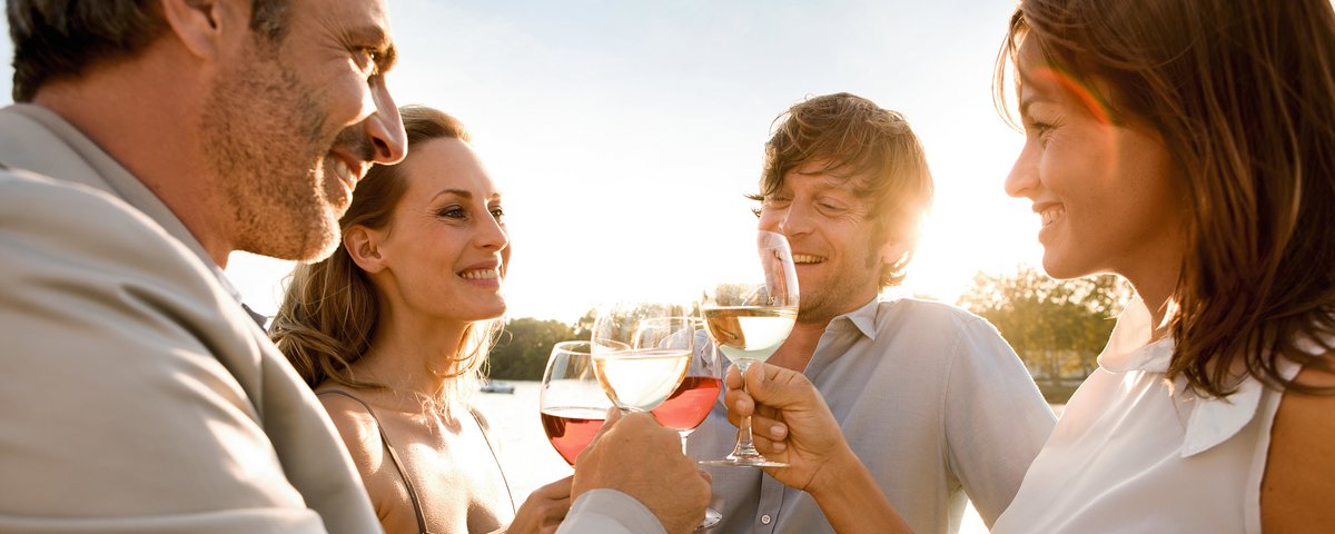 Zwei Paare stehen bei Sonnenuntergang an der Reling eines Flussschiffes, sie stoßen mit Wein an. Im Hintergrund grüne Bäume am Ufer und der Fluss.