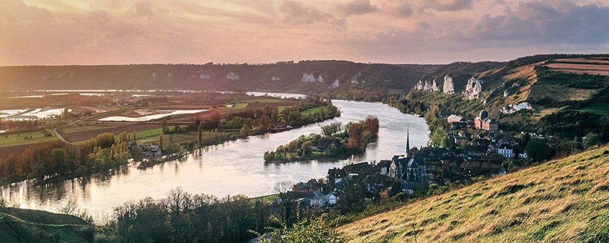 Fluss Seine, der sich durch grüne Hügel und das Dorf Les Andelys mit Kirche und Felsenlandschaft schlängelt bei Sonnenuntergang. 
