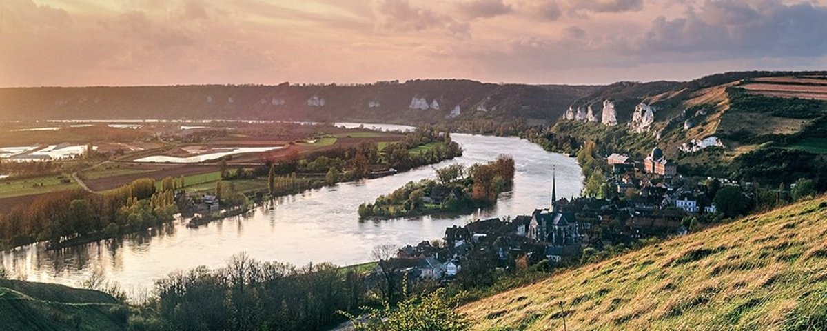 Fluss Seine, der sich durch grüne Hügel und das Dorf Les Andelys mit Kirche und Felsenlandschaft schlängelt bei Sonnenuntergang. 