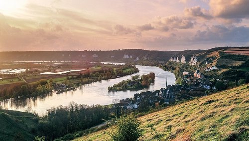 Fluss Seine, der sich durch grüne Hügel und das Dorf Les Andelys mit Kirche und Felsenlandschaft schlängelt bei Sonnenuntergang. 