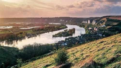 Fluss Seine, der sich durch grüne Hügel und das Dorf Les Andelys mit Kirche und Felsenlandschaft schlängelt bei Sonnenuntergang.