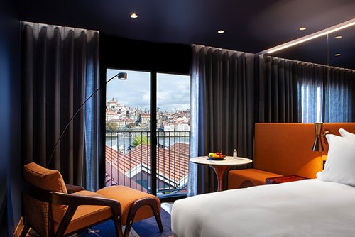 Hotelzimmer mit großem Bett, orangefarbenem Sitzbereich, Sessel und Balkon mit Blick auf Stadt und Fluss.