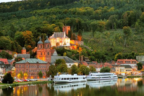 Blick auf Miltenberg mit beleuchtetem Schloss und Kirche am Flussufer, umgeben von bewaldeten Hügeln und Häusern