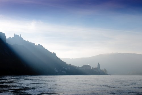 Eine mystische Flusslandschaft in der Wachau mit Hügeln am Flussufer. Am Fuss des Hügel liegt ein Dorf und auf der Spitze steht eine Burg. Die Sonne strahlt über die Hügel hinweg auf den Fluss.