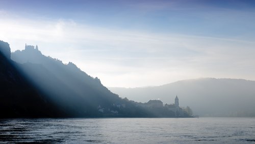 Eine mystische Flusslandschaft in der Wachau mit Hügeln am Flussufer. Am Fuss des Hügel liegt ein Dorf und auf der Spitze steht eine Burg. Die Sonne strahlt über die Hügel hinweg auf den Fluss.