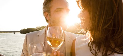 Ein Paar steht bei Sonnenuntergang mit einem Glas Wein in der Hand an der Rehling eines Flussschiffes. Im Hintergrund der Fluss und eine Uferlandschaft.