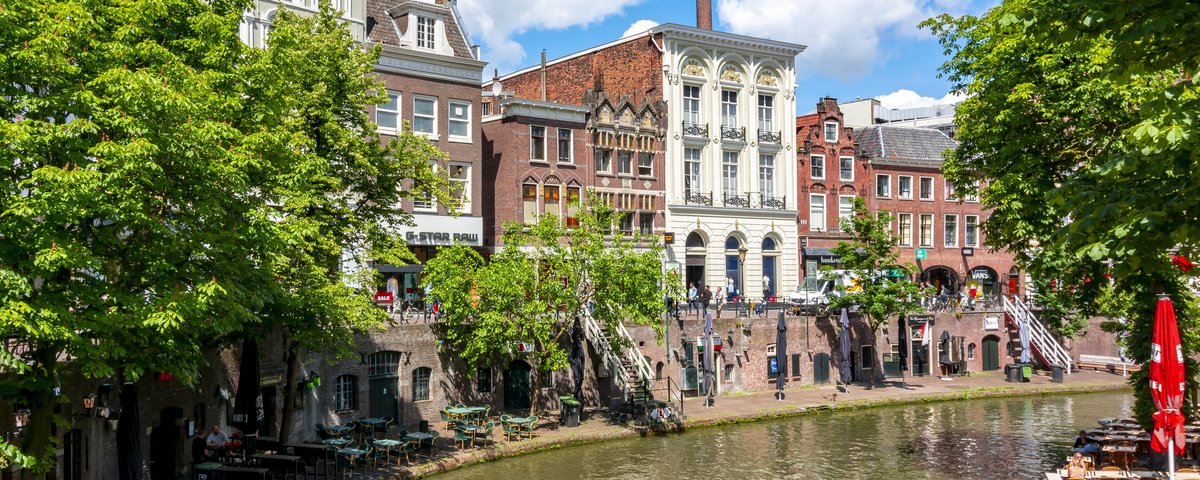 Historische Gebäude und Cafés entlang eines Kanals in Utrecht bei sonnigem Wetter mit blauem Himmel und grünen Bäumen