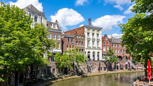 Historische Gebäude und Cafés entlang eines Kanals in Utrecht bei sonnigem Wetter mit blauem Himmel und grünen Bäumen