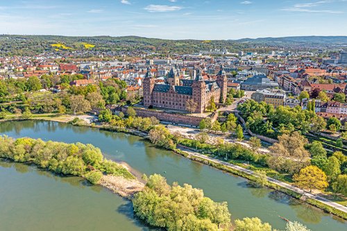Aschaffenburgs Schloss Johannisburg spiegelt sich im Main, umgeben von grünen Bäumen und einem klaren blauen Himmel.