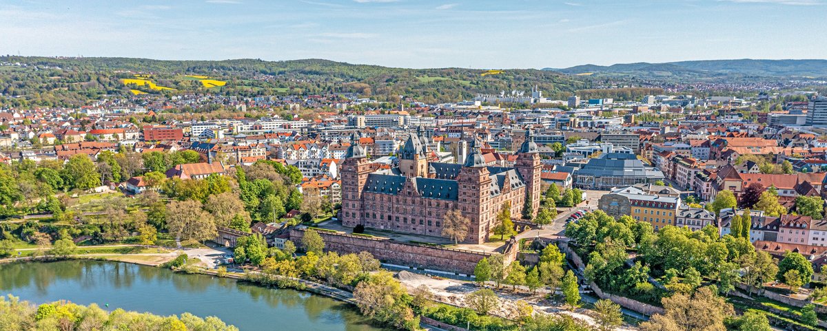 Aschaffenburgs Schloss Johannisburg spiegelt sich im Main, umgeben von grünen Bäumen und einem klaren blauen Himmel.