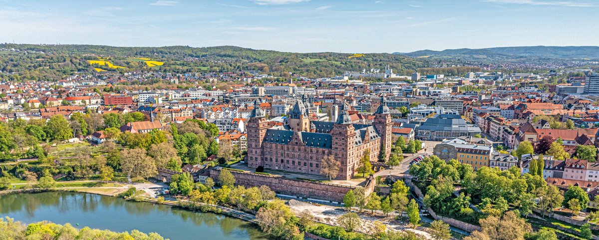 Aschaffenburgs Schloss Johannisburg spiegelt sich im Main, umgeben von grünen Bäumen und einem klaren blauen Himmel.
