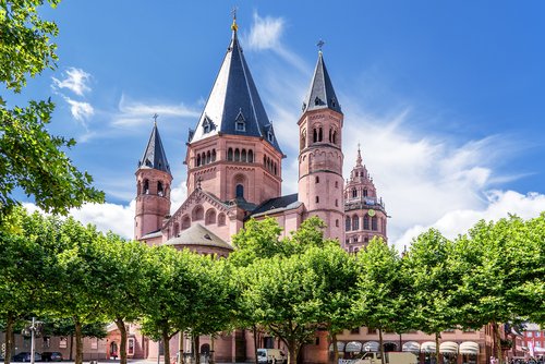 Der Mainzer Dom St. Martin mit seinen markanten roten Sandsteintürmen und spitzen Dächern, umgeben von grünen Bäumen und einem blühenden Vorgarten unter blauem Himmel