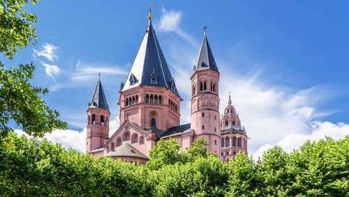 Der Mainzer Dom St. Martin mit seinen markanten roten Sandsteintürmen und spitzen Dächern, umgeben von grünen Bäumen und einem blühenden Vorgarten unter blauem Himmel