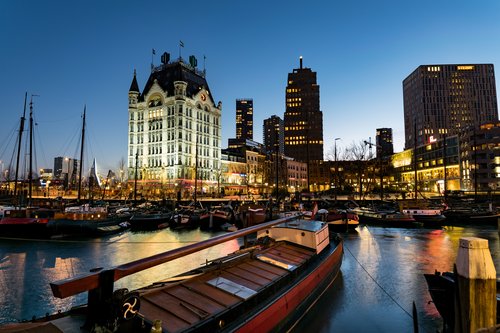 Blick aus einem kleinen Hafen in Rotterdam bei Nacht. Im Vordergrund ankern historische Boote, im Hintergrund beleuchtete Gebäude