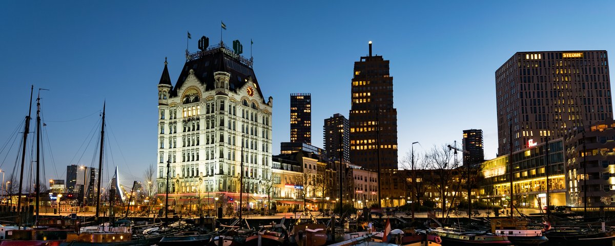 Blick aus einem kleinen Hafen in Rotterdam bei Nacht. Im Vordergrund ankern historische Boote, im Hintergrund beleuchtete Gebäude