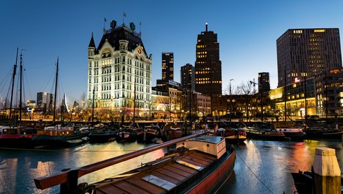 Blick aus einem kleinen Hafen in Rotterdam bei Nacht. Im Vordergrund ankern historische Boote, im Hintergrund beleuchtete Gebäude