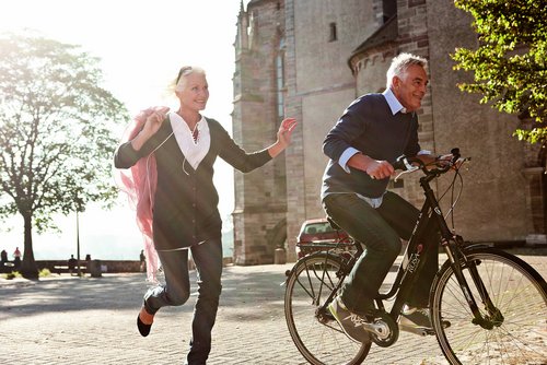 Ein Mann fährt auf einem Fahrrad von A-ROSA durch Basel bei Sonnenschein. Eine Frau läuft neben dem Fahrrad her. Im Hintergrund sind ein historisches Gebäude und ein Baum.