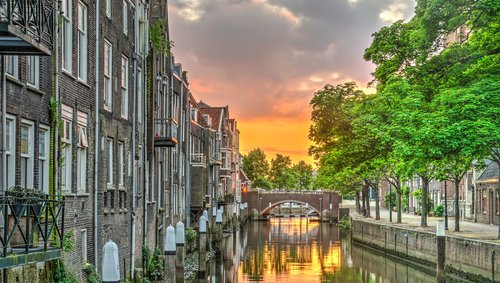 Kanal in Dordrecht mit historischen Backsteinhäusern links, grünen Bäumen rechts und einer Brücke im Hintergrund bei Sonnenuntergang