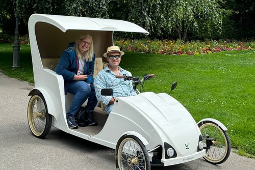 Eine Frau lässt sich mit einer Velogondel durch einen Park fahren und lächelt in die Kamera. Im Hintergrund sind ein Baum und Blumen.