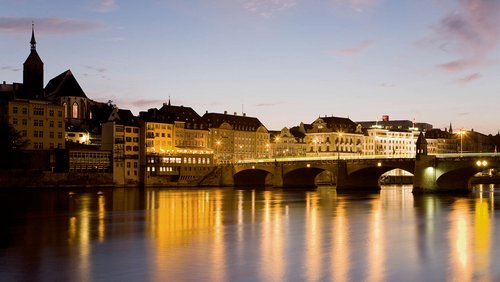 Panoramablick auf den Rhein in Basel in Abendstimmung mit historischen Gebäuden am Ufer und einer Brücke, die über den Fluss führt. Die Lichter spiegeln sich im Wasser. 