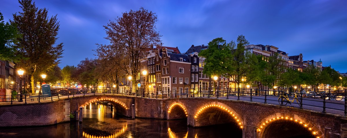 Beleuchtete Steinbrücke über einen Kanal in Amsterdam bei Abenddämmerung mit historischen Häusern und Bäumen im Hintergrund
