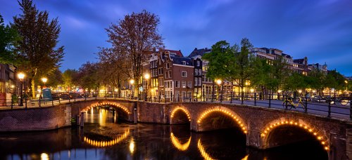 Beleuchtete Steinbrücke über einen Kanal in Amsterdam bei Abenddämmerung mit historischen Häusern und Bäumen im Hintergrund