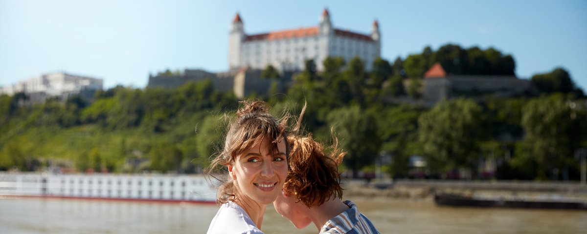 Zwei Frauen stehen an der Reling auf einem Flusskreuzfahrtschiff auf der Donau mit Blick auf ein weiteres Schiff und der Burg Bratislava auf einem bewachsenene Hügel im Hintergrund.