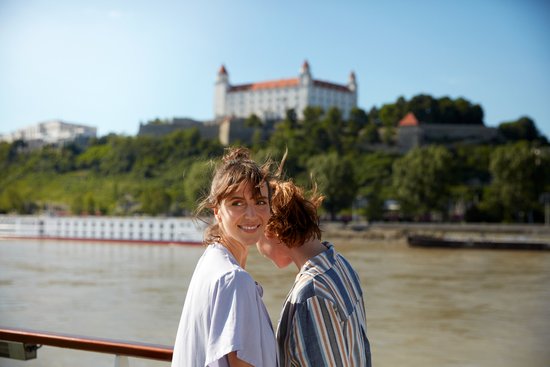 Zwei Frauen stehen an der Reling auf einem Flusskreuzfahrtschiff auf der Donau mit Blick auf ein weiteres Schiff und der Burg Bratislava auf einem bewachsenene Hügel im Hintergrund.