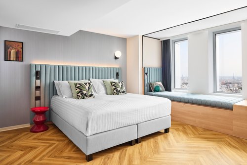 Modernes Hotelzimmer mit großem Doppelbett, gepolstertem Kopfteil in Hellblau, roten Nachttisch, Fensterbank mit Sitzkissen und Blick auf Stadt.