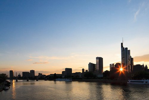 Frankfurter Skyline bei Sonnenuntergang, mit Wolkenkratzern und dem Main im Vordergrund mit Schiffen und einer Brücke.
