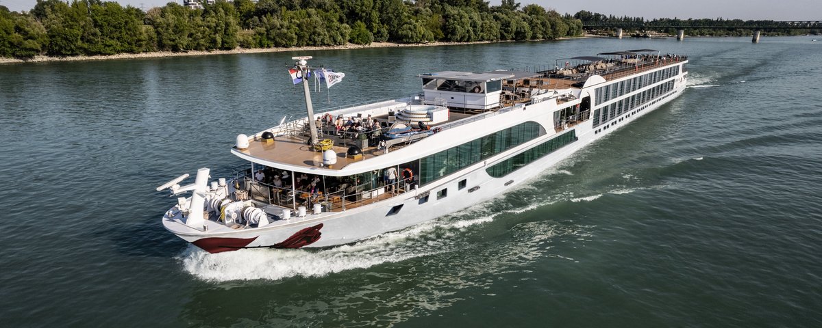 Ein Flusskreuzfahrtschiff fährt auf dem Rhein an einem bewaldeten Ufer entlang. Das Sonnendeck ist mit Whirlpool, Sonnenliegen und Sonnensegeln ausgestattet.