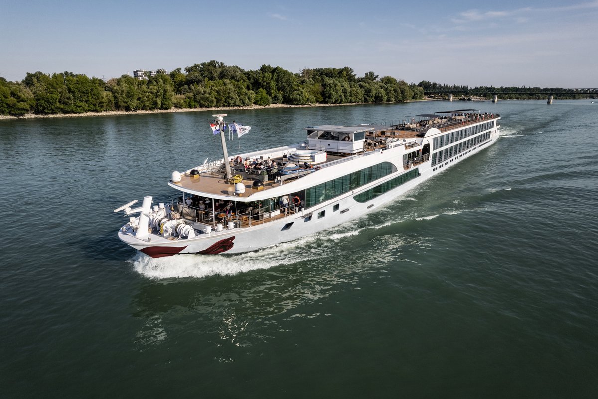 Ein Flusskreuzfahrtschiff fährt auf dem Rhein an einem bewaldeten Ufer entlang. Das Sonnendeck ist mit Whirlpool, Sonnenliegen und Sonnensegeln ausgestattet.