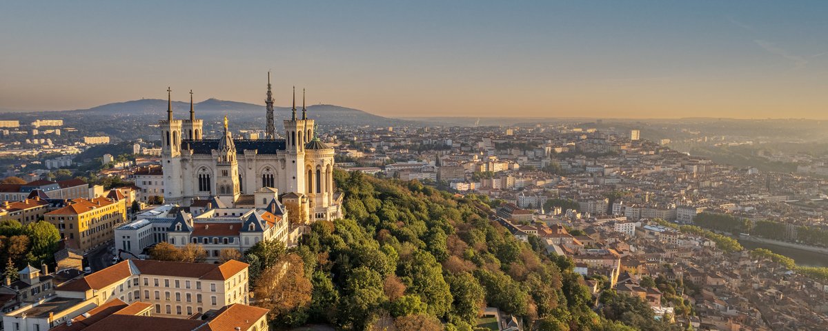 Die Basilika Notre-Dame de Fourvière in Lyon thront majestätisch auf einem Hügel, umgeben von grünen Bäumen bei Abenddämmerung. Im Hintergrund die Stadt Lyon