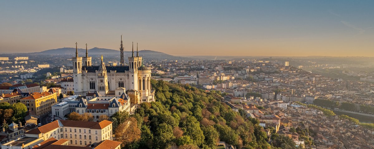 Die Basilika Notre-Dame de Fourvière in Lyon thront majestätisch auf einem Hügel, umgeben von grünen Bäumen bei Abenddämmerung. Im Hintergrund die Stadt Lyon