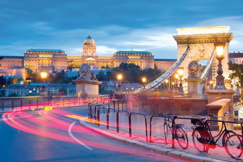 Blick auf eine beige historsiche Brücke in Budapest bei Nacht. Vorbeifahrende Autos ziehen Lichtspuren hinter sich her. Im Hintergrund stehht die Budaer Burg.