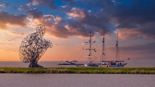 Große Drahtskulptur einer hockenden Person neben einem Segelschiff bei Sonnenuntergang am Wasser.