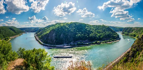 Die Loreley-Felsformation am Rhein, umgeben von grünen Hügeln und einem klaren blauen Himmel, spiegelt sich im ruhigen Flusswasser wider.