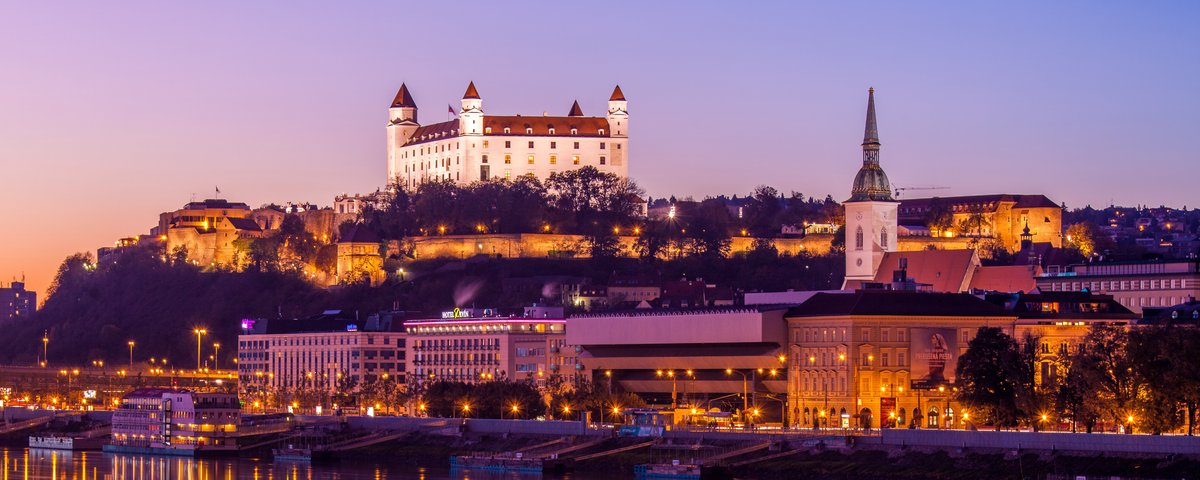 Blick auf das Donauufer bei Bratislava bei Sonnenuntergang mit der Burg Bratislava im Hintergrund, umgeben von Bäumen und der Stadt. Im Vordergrund spiegeln sich die Lichter im Fluss.