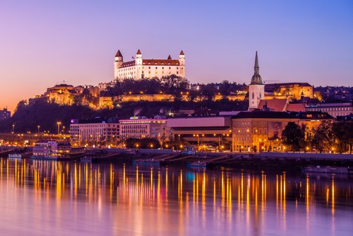 Blick auf das Donauufer bei Bratislava bei Sonnenuntergang mit der Burg Bratislava im Hintergrund, umgeben von Bäumen und der Stadt. Im Vordergrund spiegeln sich die Lichter im Fluss.