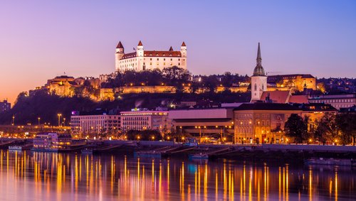 Blick auf das Donauufer bei Bratislava bei Sonnenuntergang mit der Burg Bratislava im Hintergrund, umgeben von Bäumen und der Stadt. Im Vordergrund spiegeln sich die Lichter im Fluss.