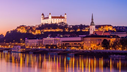 Blick auf das Donauufer bei Bratislava bei Sonnenuntergang mit der Burg Bratislava im Hintergrund, umgeben von Bäumen und der Stadt. Im Vordergrund spiegeln sich die Lichter im Fluss.