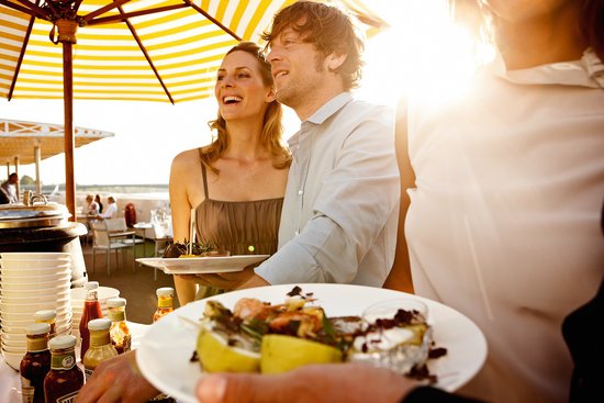 Drei Personen stehen am Buffet beim Barbecue auf dem Sonnendeck eines Flusskreuzfahrtschiffes mit Essen vom Grill, im Hintergrund der Fluss und Bäume.