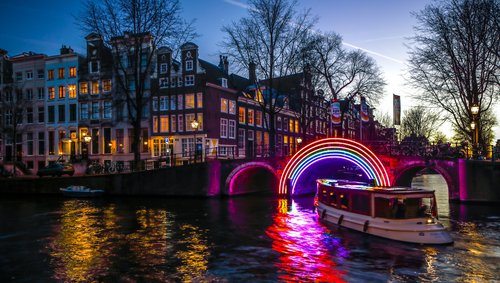 Beleuchtete Brücke mit buntem Neon-Regenbogen über einem Kanal in Amsterdam bei Dämmerung, Boot auf dem Wasser
