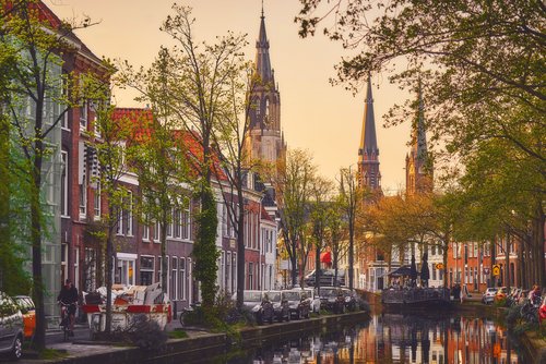 Kanal in Delft mit historischen Backsteinhäusern, Bäumen und drei hohen Kirchtürmen im Hintergrund bei Sonnenuntergang