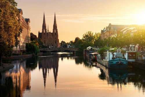Ein Fluss in Strassburg mit Booten am Ufer, umgeben von historischen Gebäuden und Bäumen mit Blick auf eine Brücke vor der Paulskirche bei Sonnenuntergang.